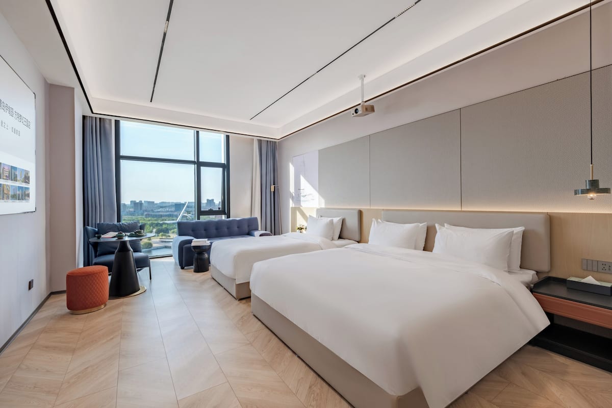 Habitación de hotel moderna con dos camas, un sofá, una mesita y un amplio ventanal con vistas a la ciudad.