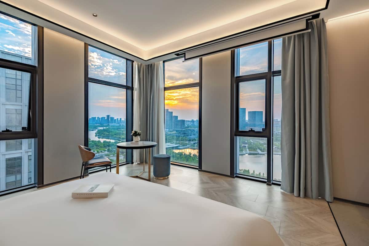 Habitación de hotel moderna con ventanas amplias, una cama, una pequeña mesa y una silla.
