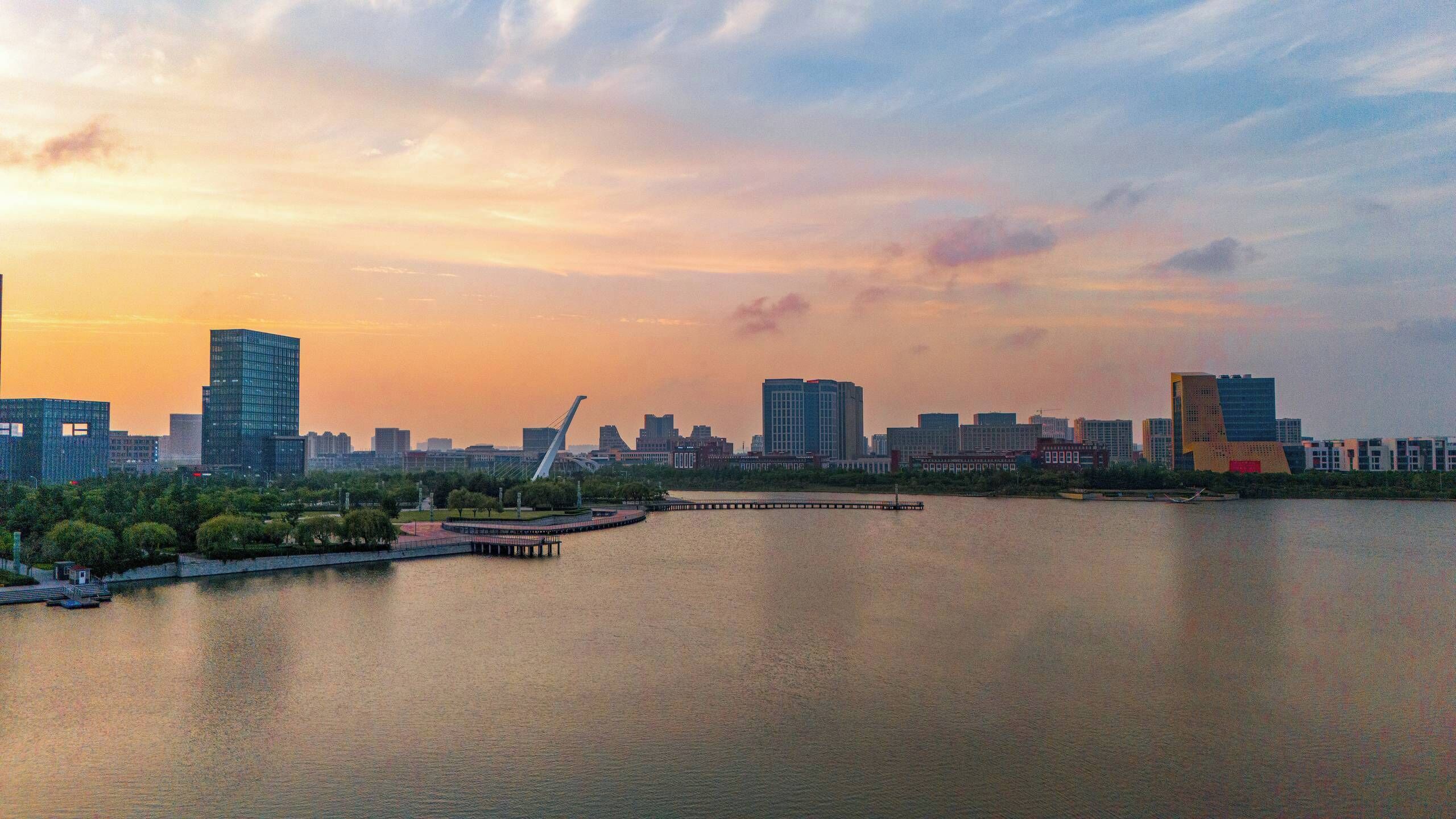 A cityscape with modern buildings, a bridge, and a body of water during sunset.