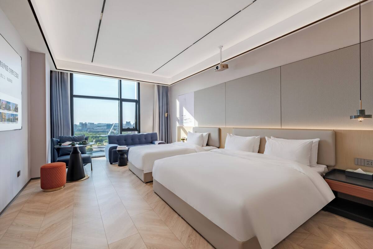 A modern hotel room with two beds, a sofa, a small table, and a large window offering a city view.