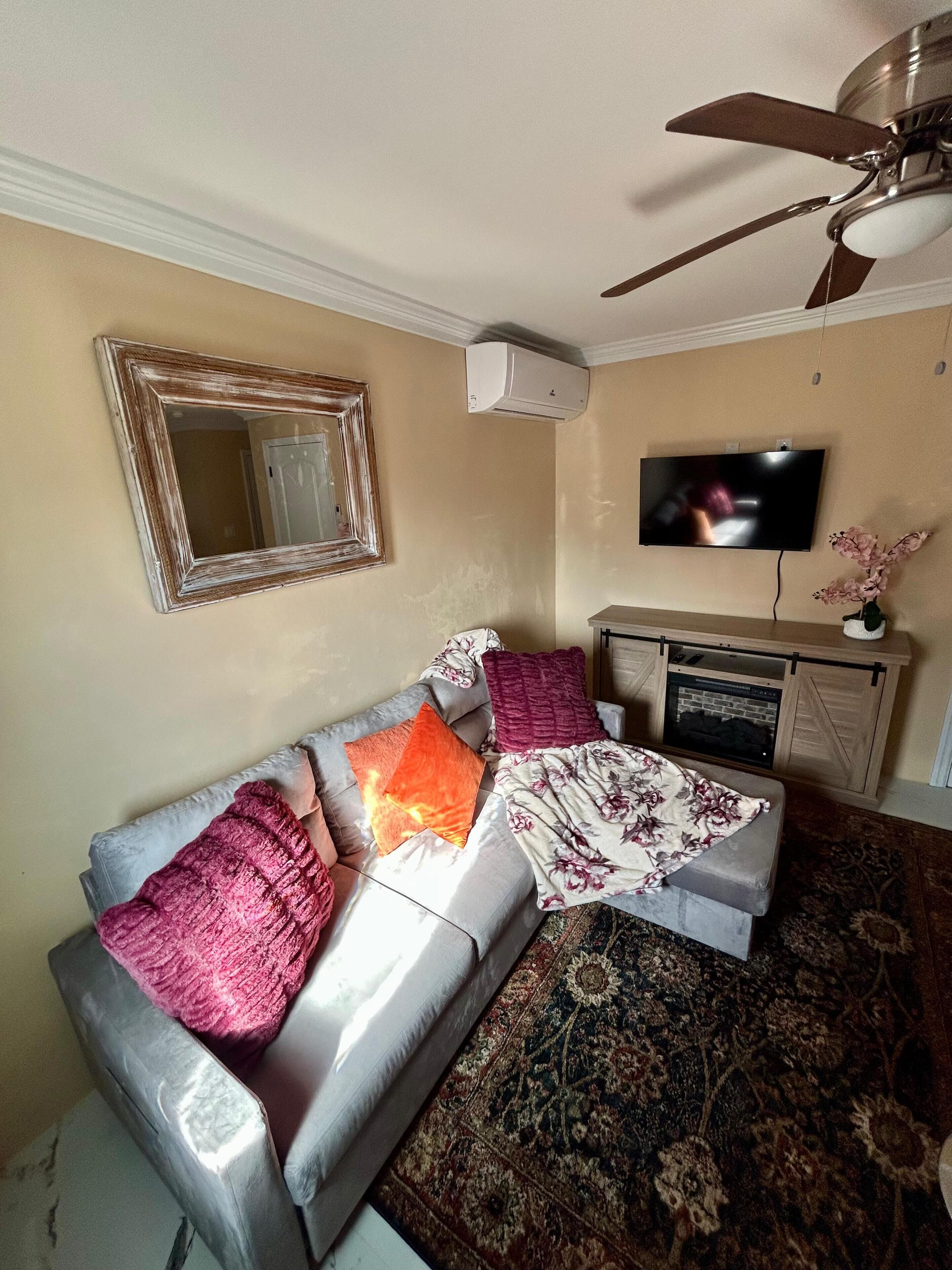 A living room with a sofa, pillows, a television, a ceiling fan, and a wall-mounted air conditioner.