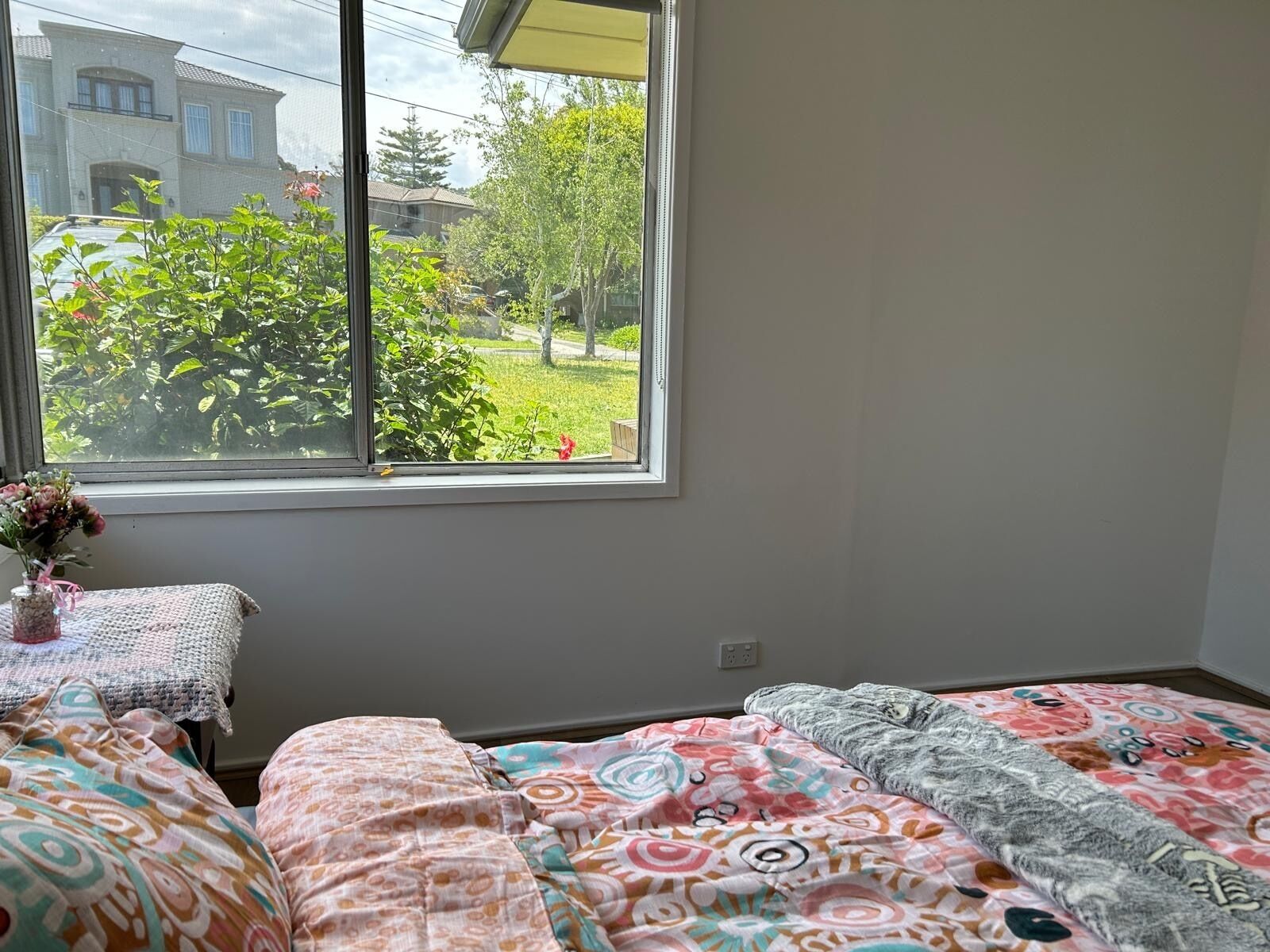 A bedroom with a large window, a small table with a vase of flowers, and two beds with patterned bedding.