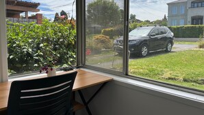 Quarto com uma grande janela com vista para uma rua residencial, uma escrivaninha de madeira e uma cadeira preta.