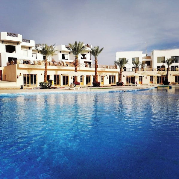 A swimming pool with a clear blue water surface, surrounded by white buildings with balconies and palm trees. - Palma Di Sharm Resort (Sharm El Sheikh)