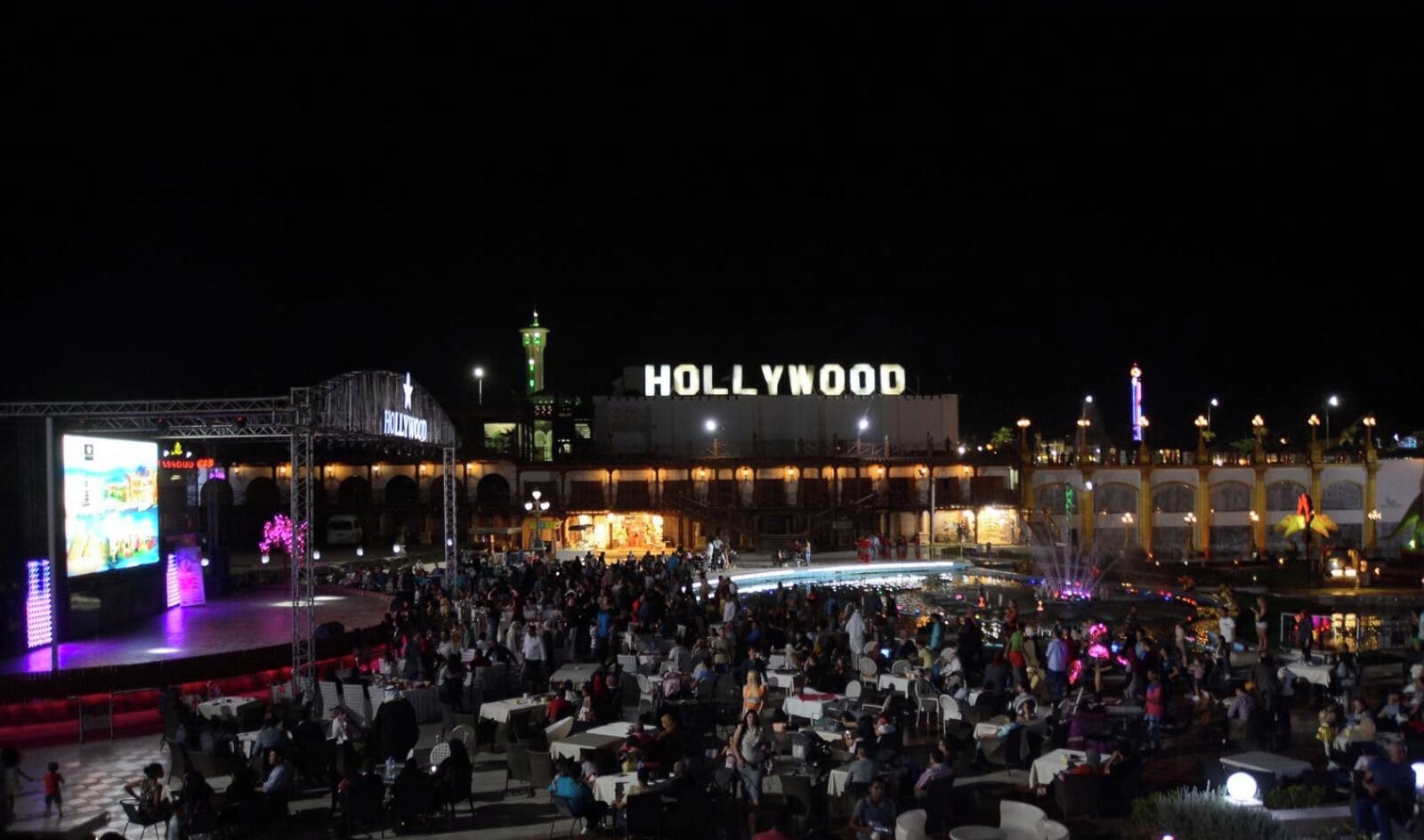 Un grande evento all'aperto con un palco e un pubblico di fronte a un edificio con la scritta 'HOLLYWOOD'.