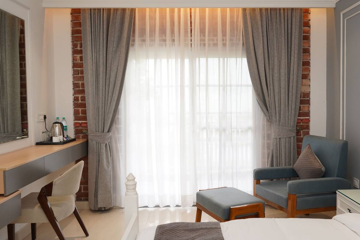 A hotel room with a bed, a desk, a chair, and a small table. There is a window with curtains and a brick wall.