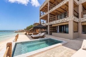 Ein Poolbereich mit Liegestühlen und Sonnenschirmen, mit Blick auf den Strand, sowie ein Gebäude mit einer steinernen Fassade.