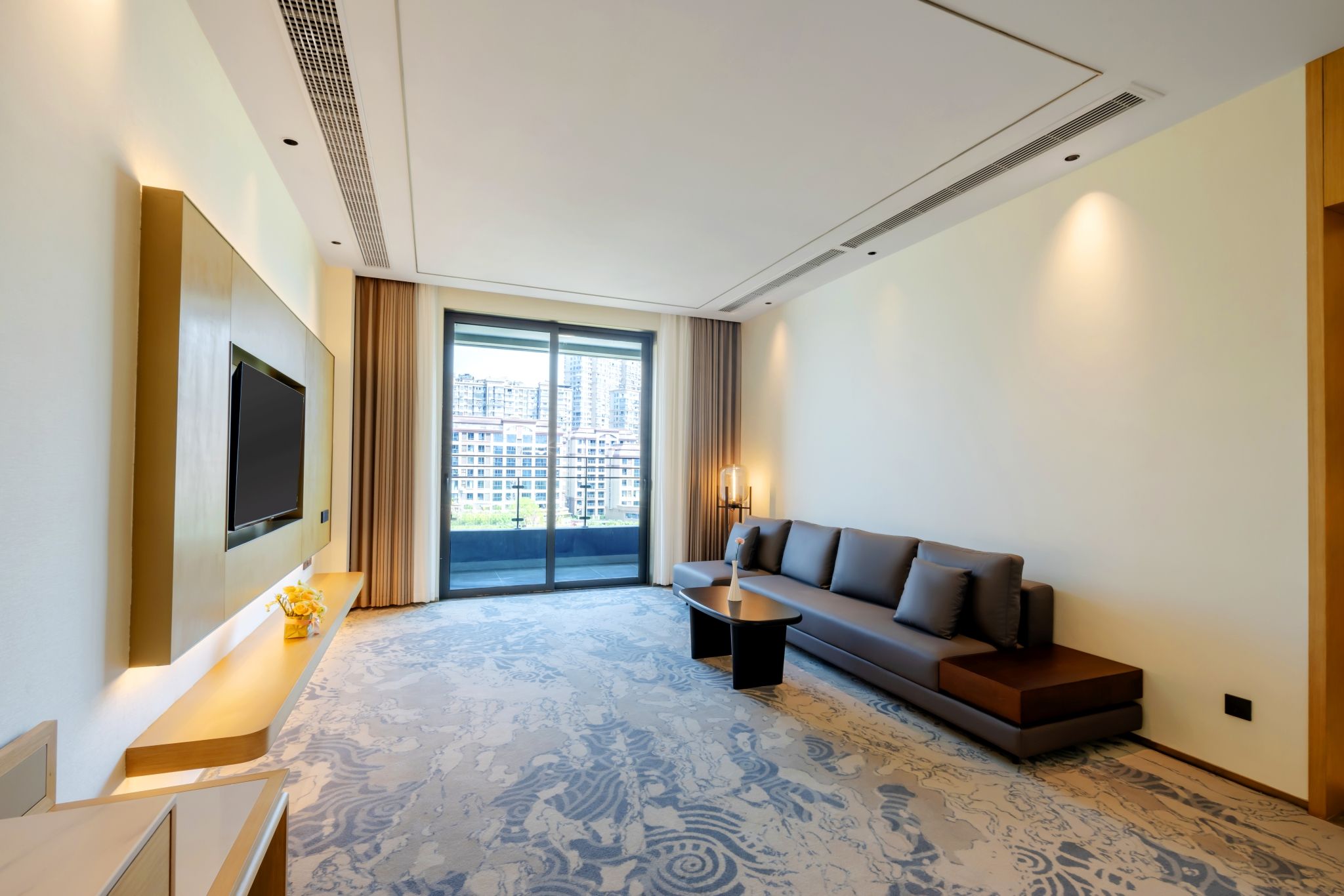A modern hotel room with a large window, a flat-screen TV mounted on the wall, a sofa, a coffee table, and a patterned carpet.