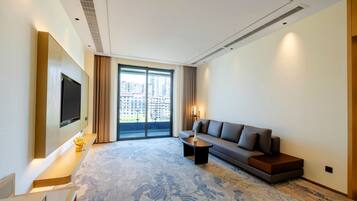 A modern hotel room with a large window, a flat-screen TV mounted on the wall, a sofa, a coffee table, and a patterned carpet.
