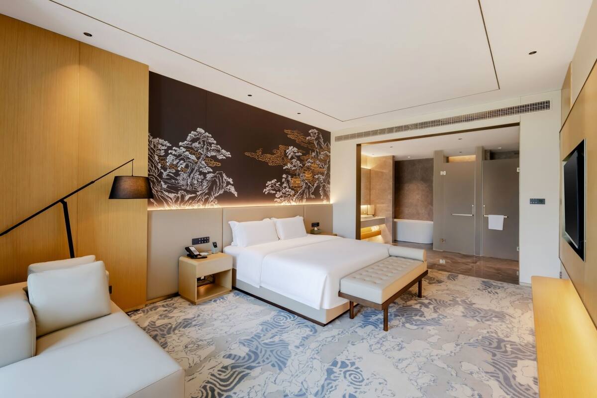 Una habitación de hotel moderna con una cama grande, mesitas de noche, un sofá, una pared decorativa y un baño visible a través de una puerta abierta.