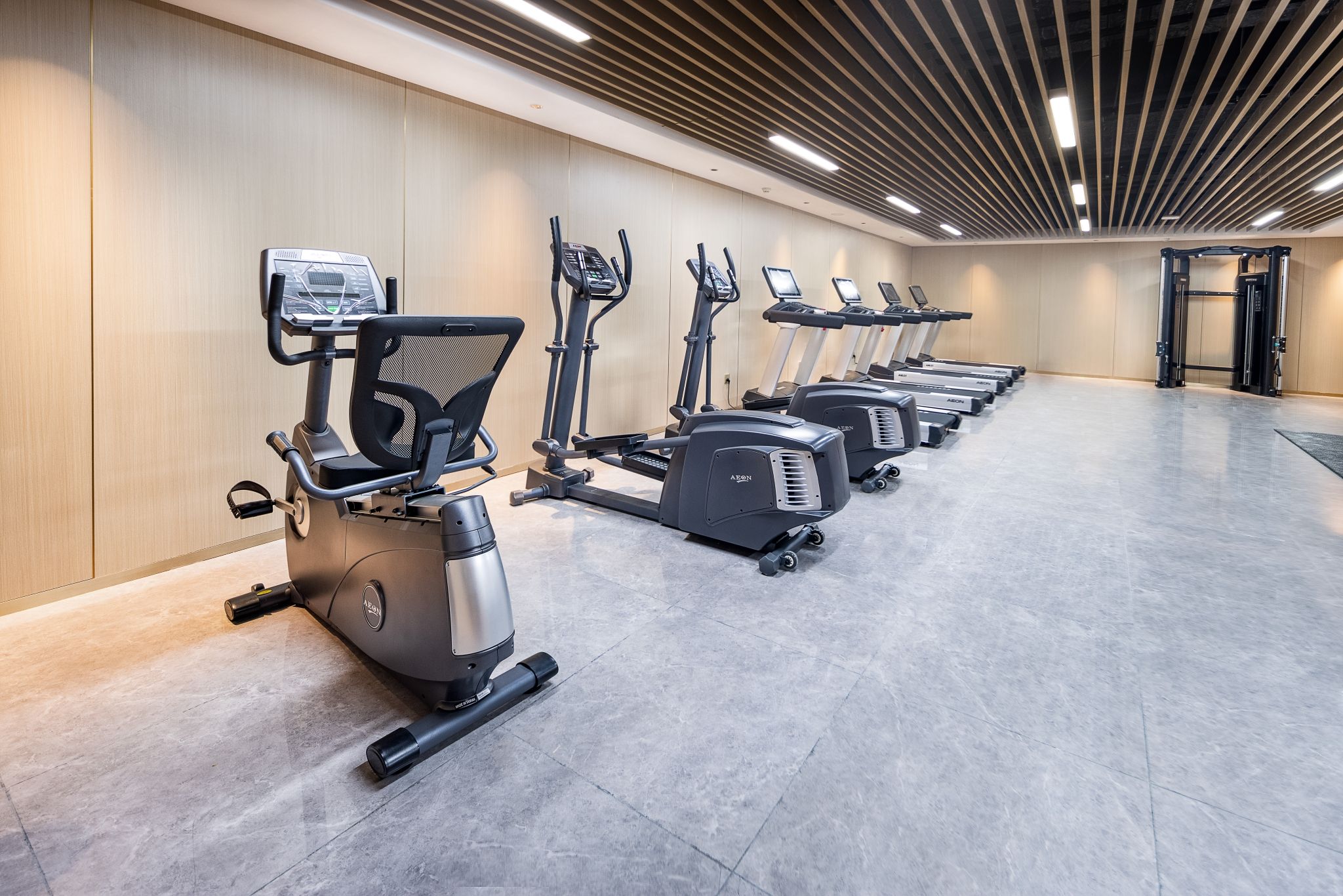 Fila de bicicletas estáticas y cintas de correr en un gimnasio con pared de madera e iluminación lineal en el techo.