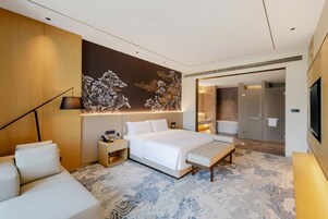 Una habitación de hotel moderna con una cama grande, mesitas de noche, un sofá, una pared decorativa y un baño visible a través de una puerta abierta.