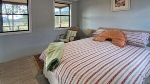 Een slaapkamer met een bed, twee ramen, een stoel en een foto aan de muur.