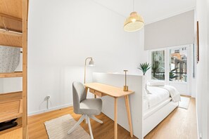 A modern, well-lit bedroom with a wooden desk, a grey chair, a bedside table, a white bed, and a view of the outside through a large window. - GuestReady - Comfort in the heart of Porto City (Porto)