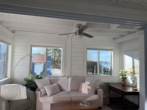 A room with a white wooden ceiling, a ceiling fan, a beige sofa with striped cushions, a wooden coffee table, and a view of a body of water through large windows. - 3-bedroom cottage in Harpswell with waterfront access to ash cove (Harpswell)