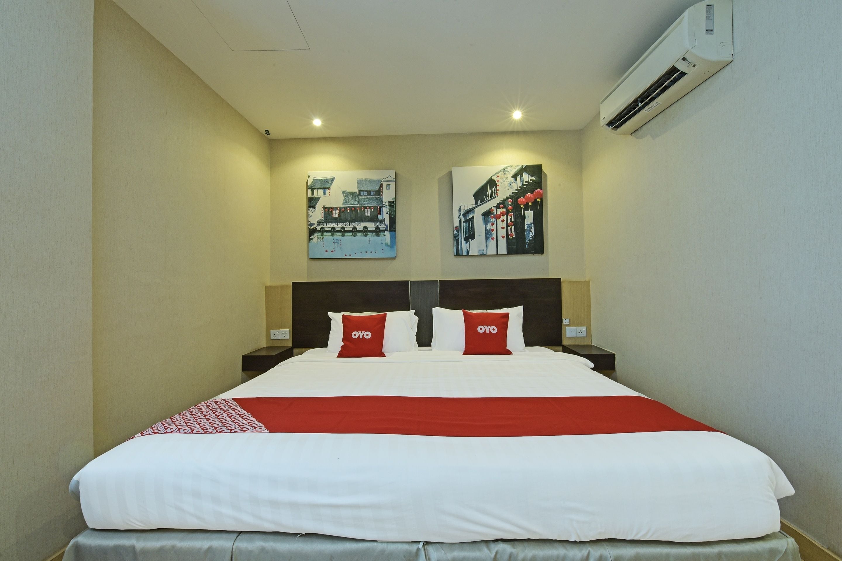 Hotelkamer met groot bed, twee rode kussens, een rood-witte plaid, twee aan de muur bevestigde schilderijen, een nachtkastje en een airconditioner.
