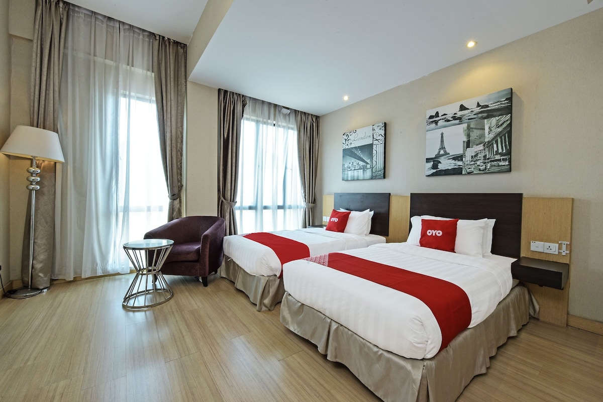 A hotel room with two beds, a chair, a lamp, and artwork on the wall.
