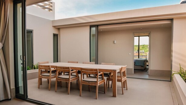Un comedor moderno con una mesa rectangular y seis sillas de madera, un divisor corredero de vidrio que da a un balcón y vistas a una zona residencial.