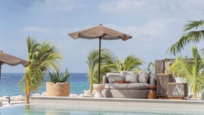Zona de piscina con un sofá y dos sombrillas tejidas, rodeada de palmeras y vistas al océano.