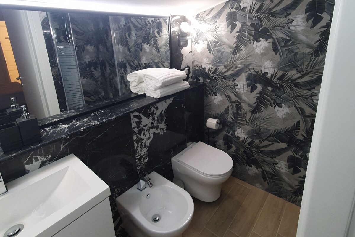 A compact bathroom with a black and white floral wallpaper, a white sink, a bidet, and a toilet.
