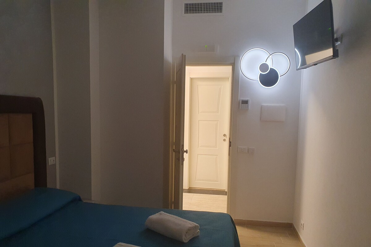 A hotel room with a bed, a flat-screen TV mounted on the wall, a door leading to another room, and a light fixture above the door.
