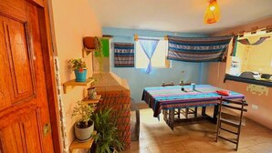 Une pièce chaleureuse avec une porte en bois, une petite table recouverte d’une nappe colorée, un banc et un luminaire suspendu.