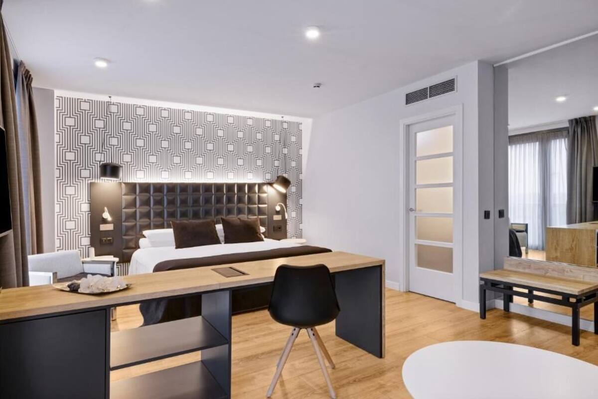 A modern hotel room with a bed, desk, and chair, a patterned wall, and a door with frosted glass panels.