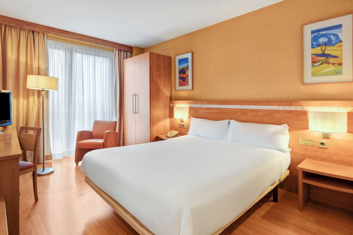 A hotel room with a large bed, a desk with a lamp, a chair, a wardrobe, and a painting on the wall.