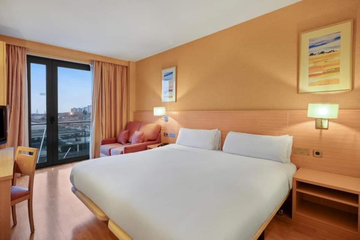 A hotel room with a large bed, a desk, a chair, a sofa, and a view of the city through a large window.