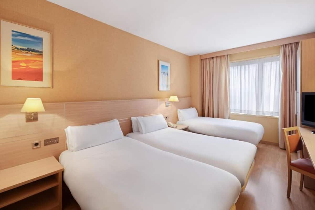 A hotel room with three beds, a desk, a chair, a TV, and two wall-mounted lamps.