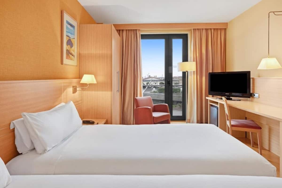 A hotel room with a large bed, a desk with a television, a chair, and a view of a cityscape through the window.