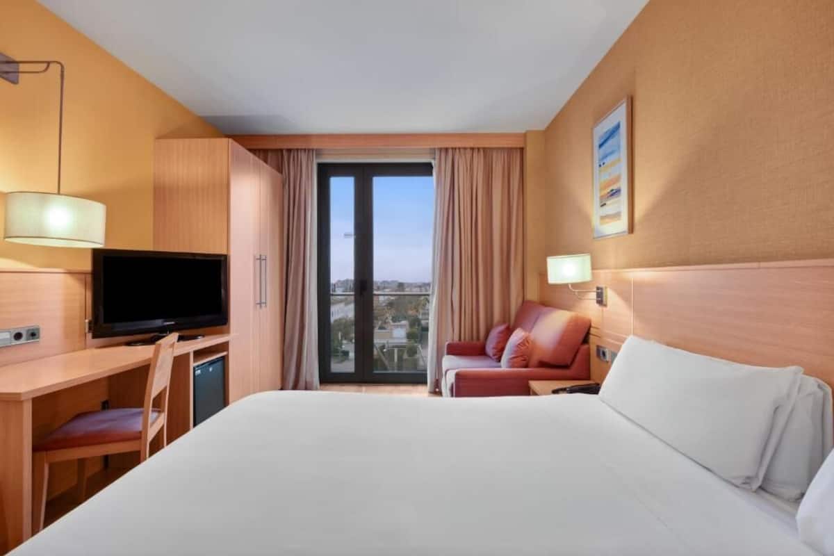 A hotel room with a bed, a desk with a TV, a chair, a sofa, and a view of the city through the window.
