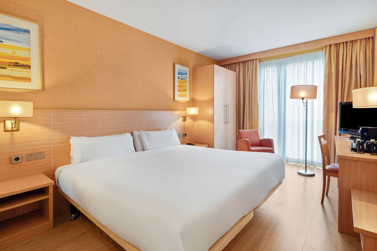 A hotel room with a large bed, a desk with a chair, a TV, and a window with curtains.