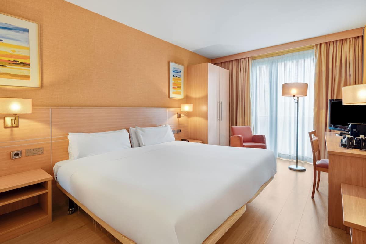 A hotel room with a large bed, a desk with a chair, a TV, and a window with curtains.