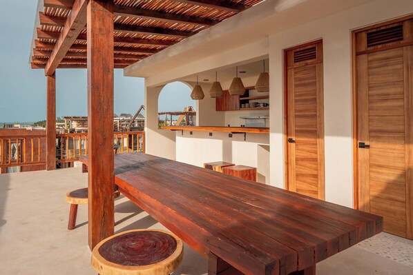 Una terraza de madera con una mesa larga y taburetes, una zona de cocina con luces colgantes, y vista al mar.