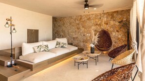 Un salon moderne avec un mur en pierre, un canapé, deux fauteuils en osier, une lampe sur pied et un ventilateur de plafond.