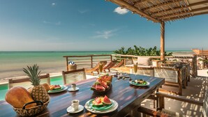 A wooden table set with plates of watermelon and pineapple, a basket of bread, and cups of coffee, overlooking a beach with a clear sky. - Hotel Boutique Casa Muuch Holbox - Solo Adultos (Isla Holbox)