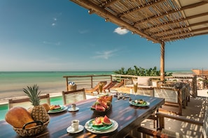 A wooden table set with plates of watermelon and pineapple, a basket of bread, and cups of coffee, overlooking a beach with a clear sky. - Hotel Boutique Casa Muuch Holbox - Solo Adultos (Isla Holbox)
