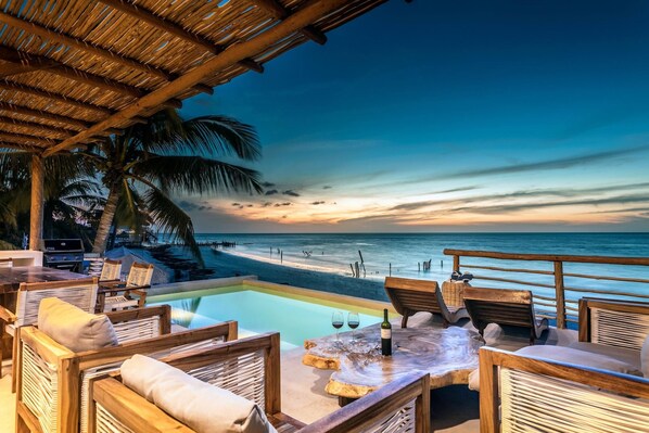 Eine Strandlounge mit Pool, Holzmöbeln und Blick auf den Sonnenuntergang über dem Meer.