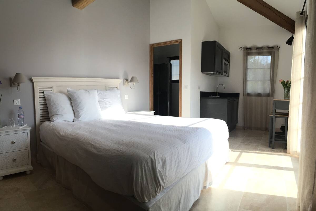 A bedroom with a large bed, bedside tables, a nightstand, a wall-mounted TV, a window with curtains, and a small bathroom area with a sink and mirror.