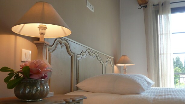 A bedroom with a bed, two bedside lamps, a nightstand with a vase of flowers, and a window with curtains. - Etoile De Saint Paul (Saint-Paul-de-Vence)