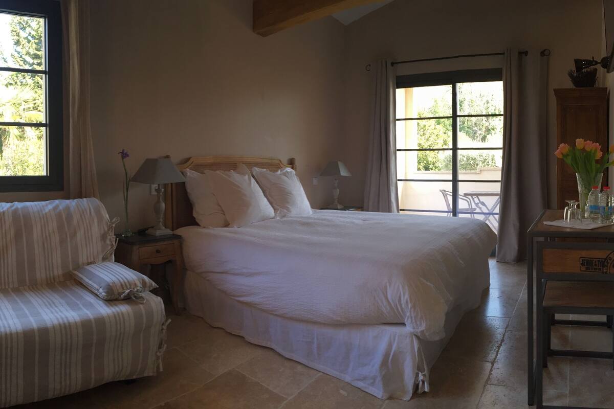 A bedroom with a bed, a sofa, a nightstand, a lamp, and a window with a view of greenery.