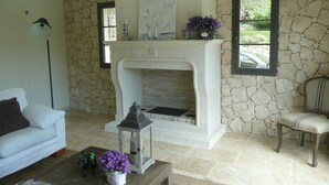 A living room with a stone wall, a fireplace, a white sofa, a chair, and a coffee table with a magazine and flowers. - Etoile De Saint Paul (Saint-Paul-de-Vence)