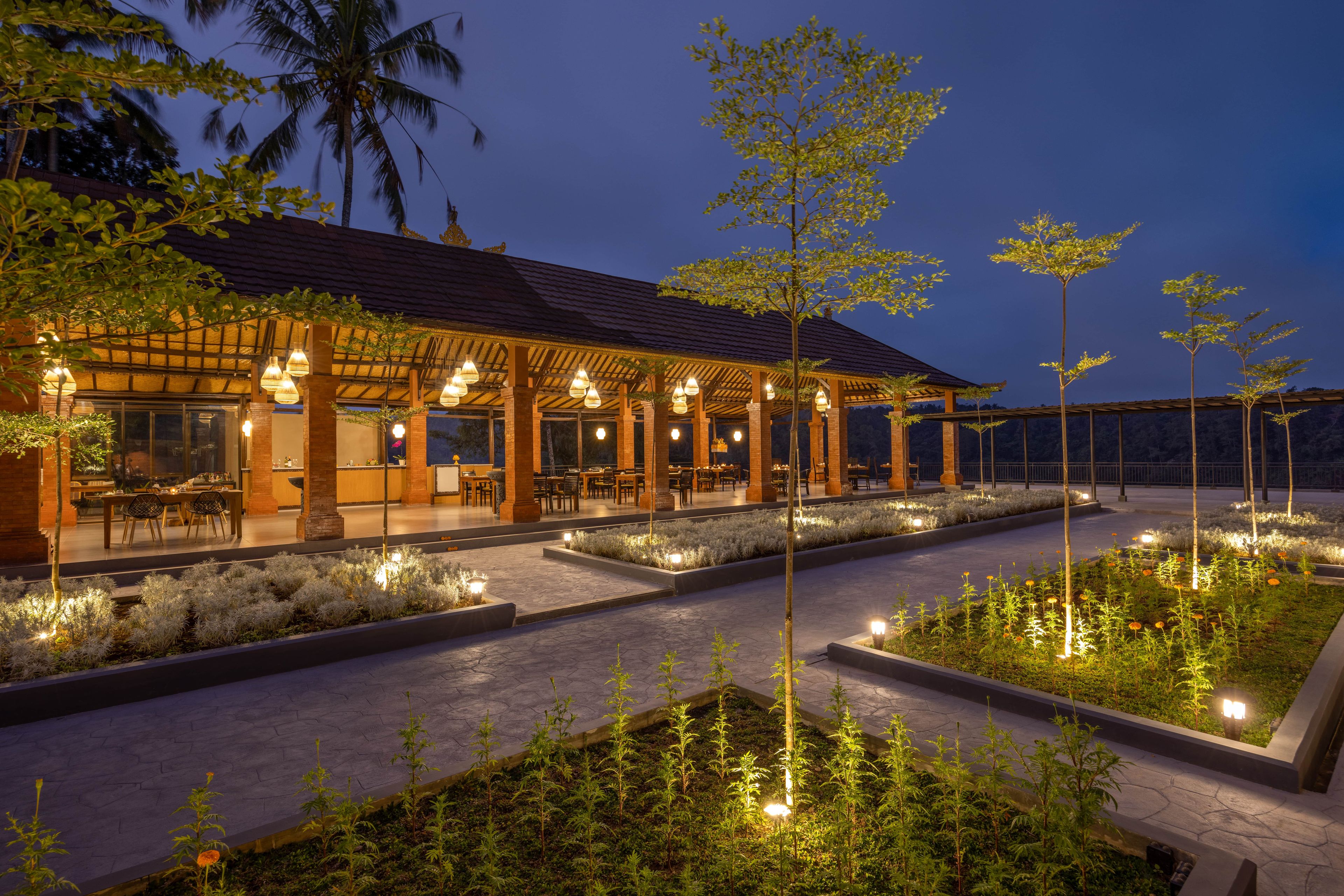 A well-lit outdoor dining area with tables and chairs, surrounded by landscaped gardens and trees.