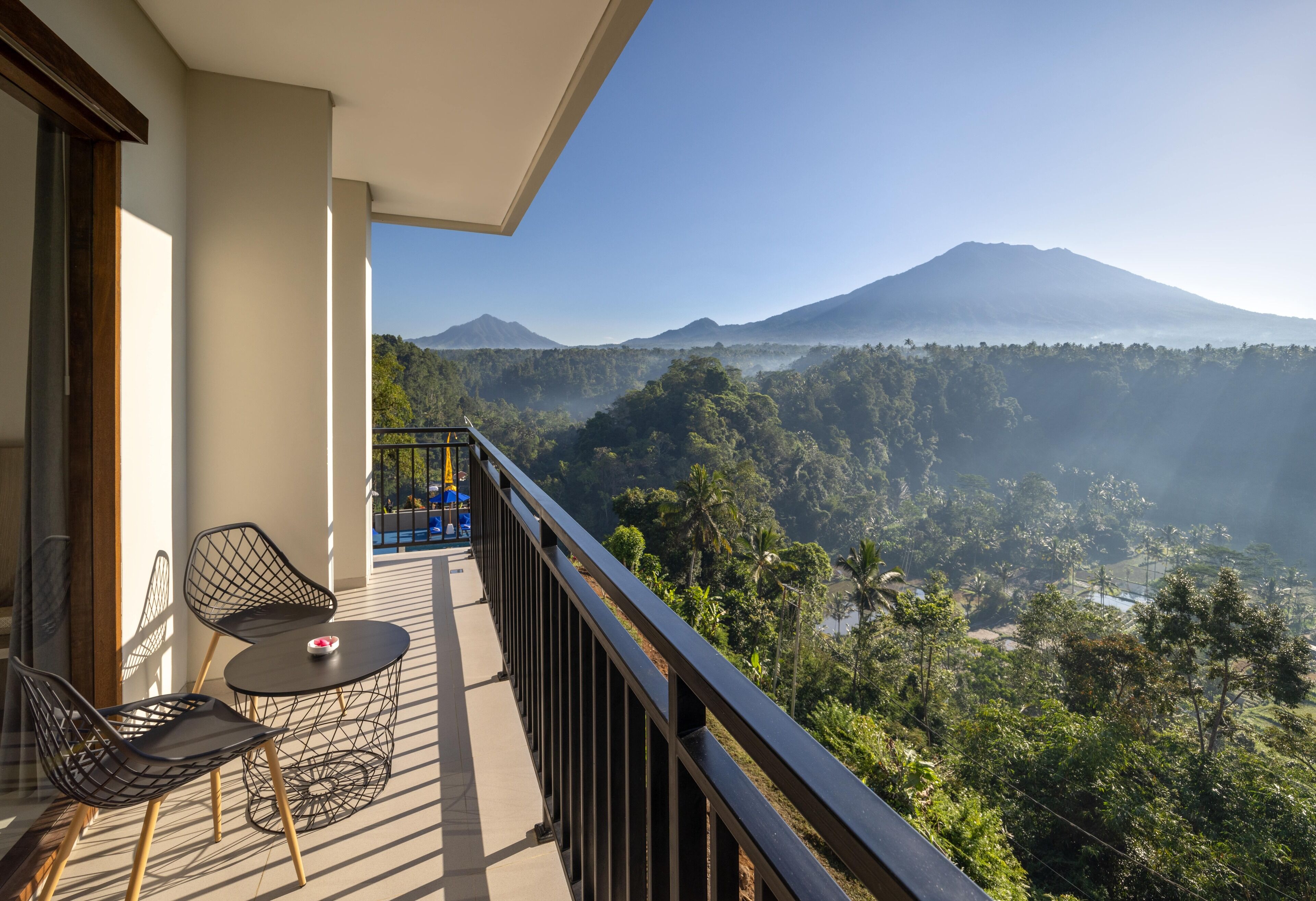 A balcony with two chairs and a table, overlooking a mountainous landscape with a view of a volcano and lush greenery.