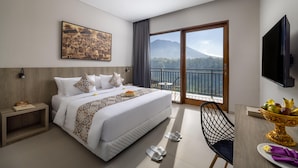 A hotel room with a large bed, a desk with a fruit bowl, a TV, and a balcony with a mountain view.