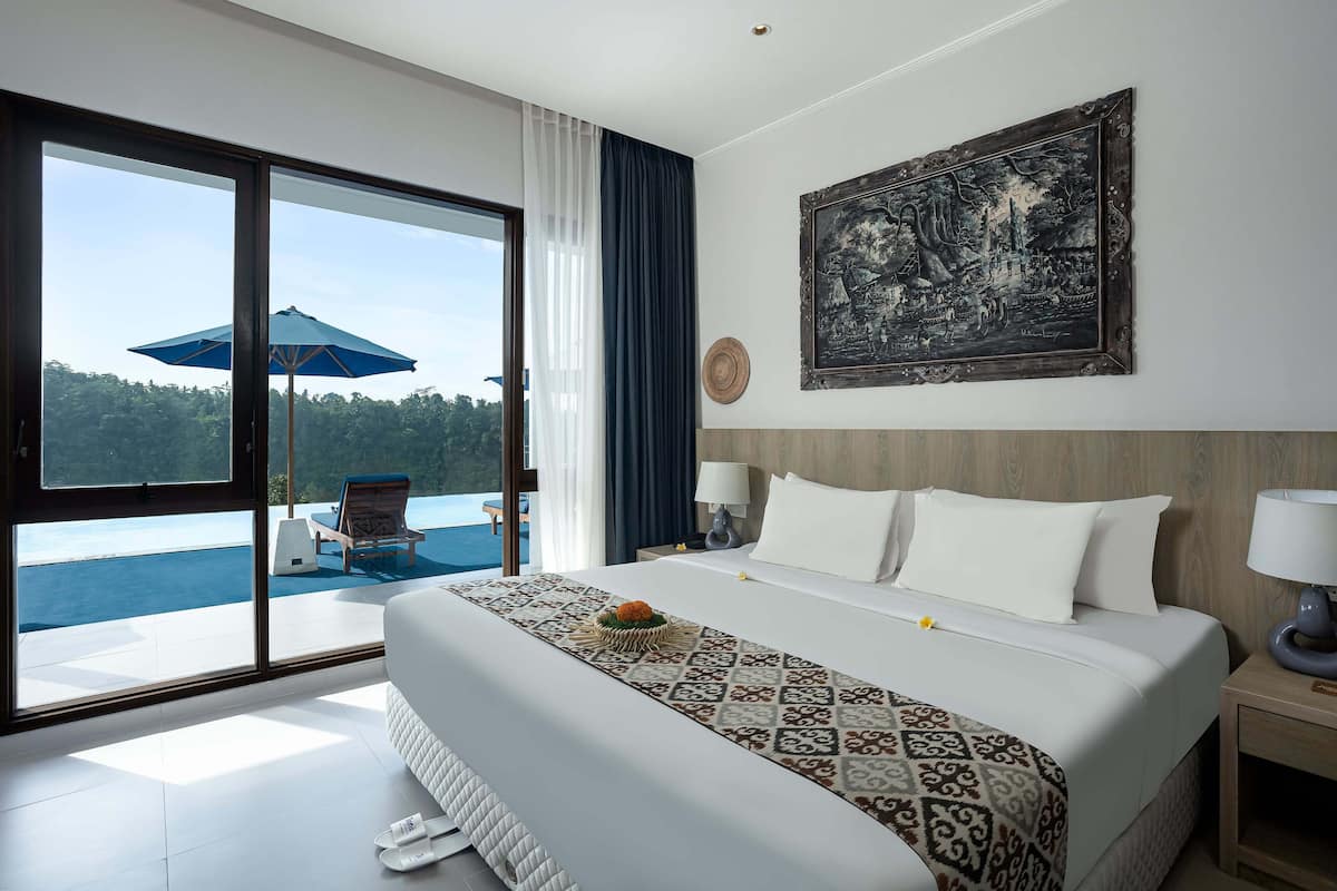 A hotel room with a large bed, a view of the sea, a painting on the wall, and a balcony with a chair and umbrella.