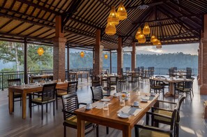 Un restaurante con mesas y sillas de madera, lámparas de mimbre colgantes y vistas a un paisaje montañoso.