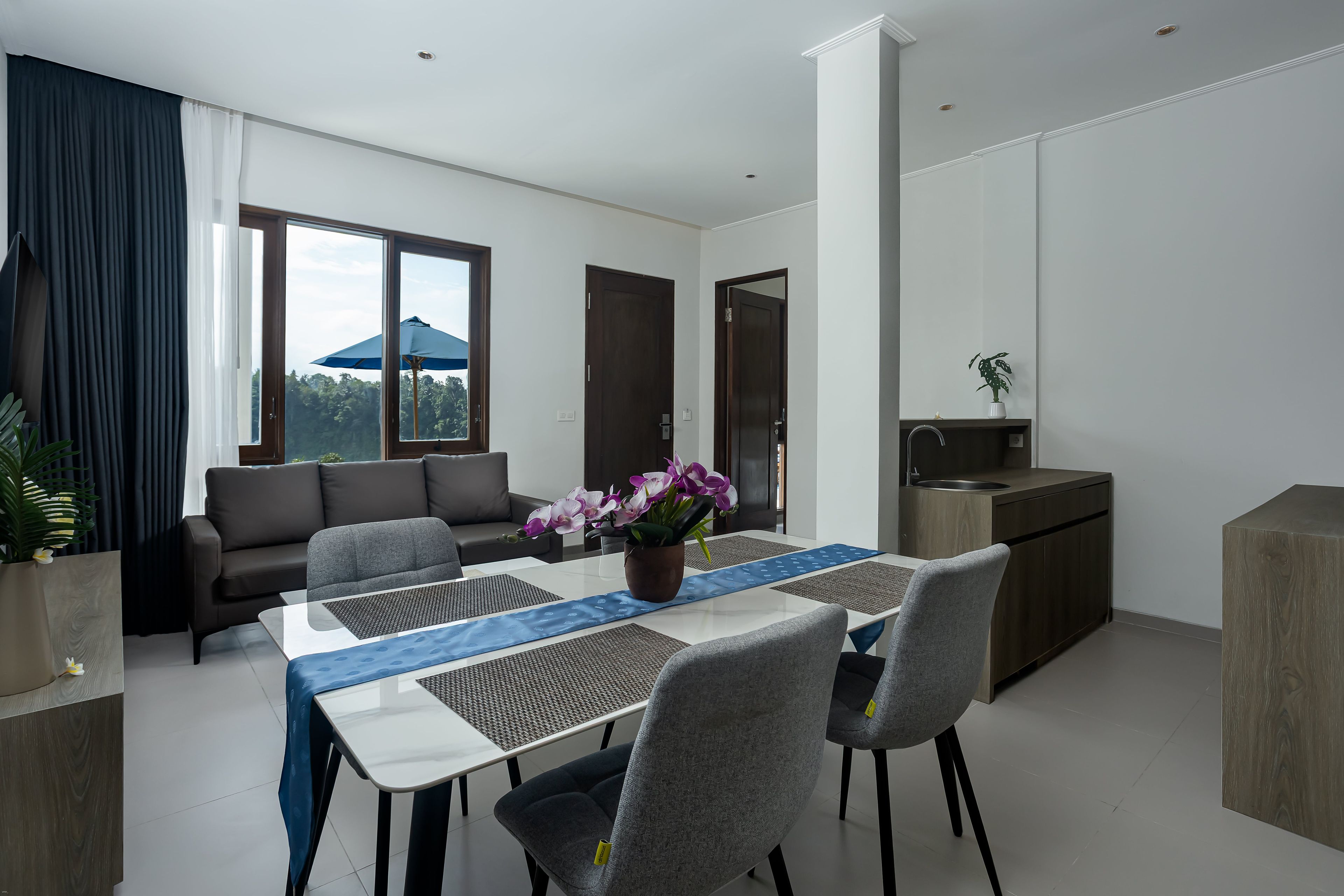 A modern living room with a sofa, a dining table with chairs, a kitchen area, and a large window with a view of greenery.