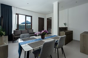 A modern living room with a sofa, a dining table with chairs, a kitchen area, and a large window with a view of greenery. - Tapa Agung View Hotel & Restaurant (Rendang)